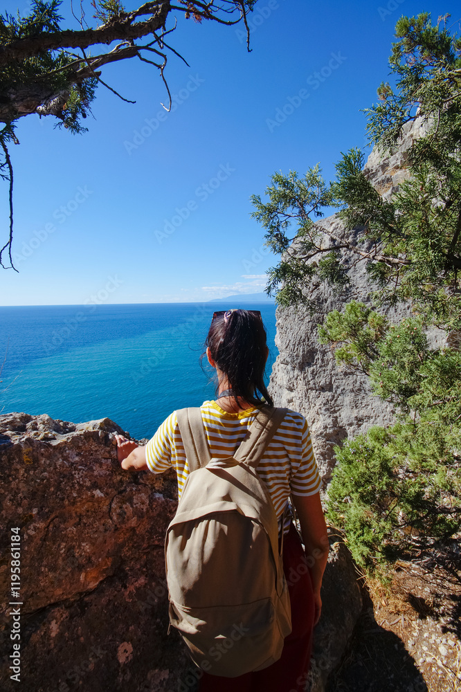 A young female traveler with a backpack sits on the top of Karaul-Oba mountain, Novy Svet village, Crimea, enjoying the sea view. A tourist with a backpack travels through the Crimea