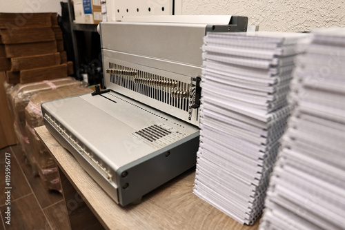 Modern binding machine and stacks of notebooks on wooden table indoors