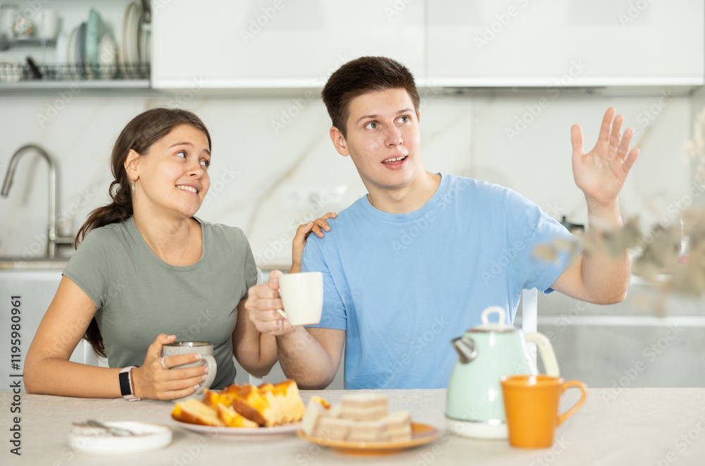 Positive young man and woman communicating merrily in the kitchen while having tea with pie