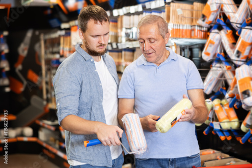 Senior man and guy carefully examines bristles of paint brush and painting roller in paintwork material department of construction store. Shop of paint and varnish materials in supermarket of building