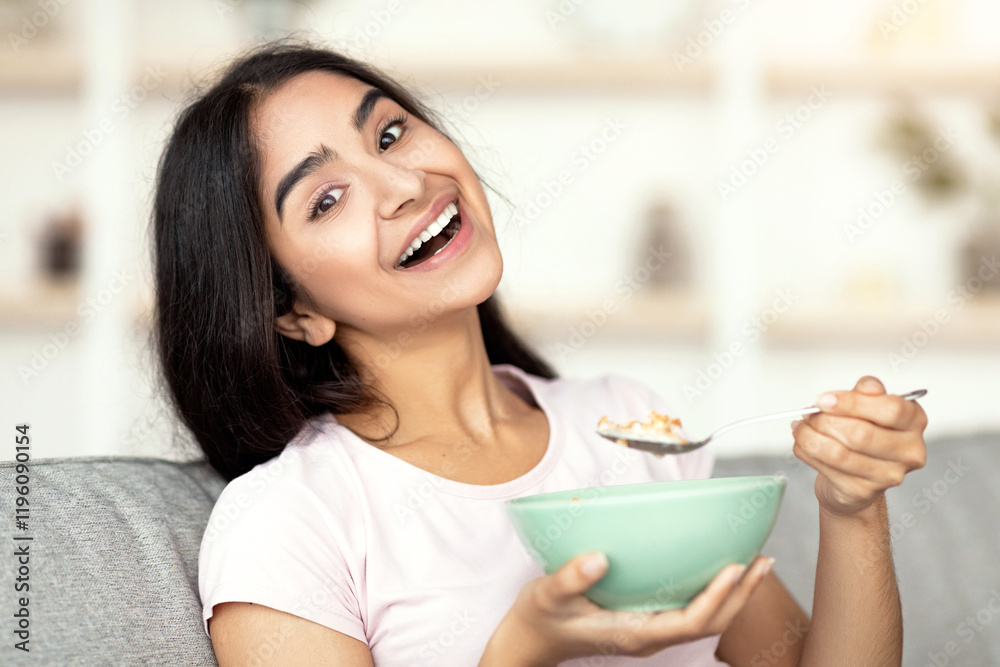 Joyful Indian lady eating granola or cereal with milk on comfy couch at home, smiling at camera. Lovely Eastern woman leading healthy lifestyle, having wholesome meal. Balanced nutrition concept Joyful Indian lady eating granola or cereal with milk on comfy couch at home, smiling at camera. Lovely Eastern woman leading healthy lifestyle, having wholesome meal. Balanced nutrition concept