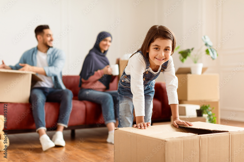 Happy kid daughter pushing carton box on floor, girl excited about moving day or relocation, cheerful child playing unpacking in new home, mom and dad sitting on sofa having fun packing together Happy kid daughter pushing carton box on floor, girl excited about moving day or relocation, cheerful child playing unpacking in new home, mom and dad sitting on sofa having fun packing together