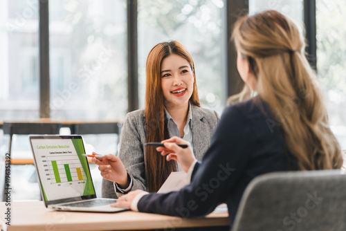 Two businesswomen discussing carbon emission reduction strategies with laptop displaying graph in modern office setting, fostering collaboration and sustainability