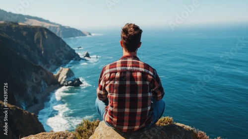 Individual enjoys a peaceful moment while sitting on a rocky cliff, gazing at the vast ocean and beautiful coastline under clear blue skies during midday