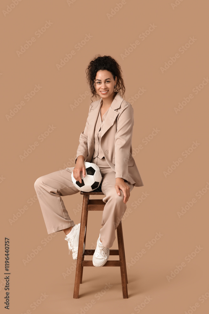 Fototapeta premium Female African-American soccer trainer in business suit with ball sitting on chair against beige background