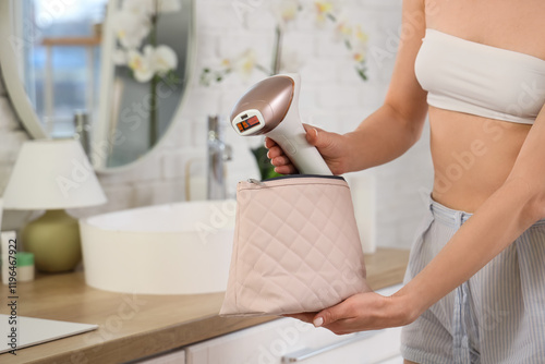 Beautiful young woman with modern photoepilator and cosmetic bag in bathroom, closeup photo