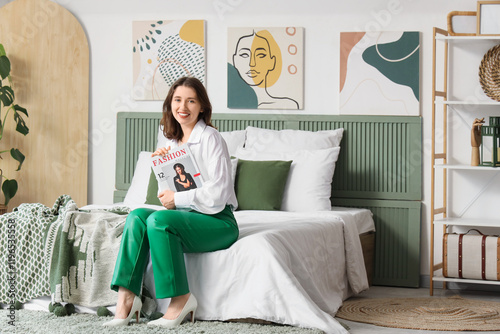 Young woman with magazine sitting near stylish paintings on light wall in bedroom