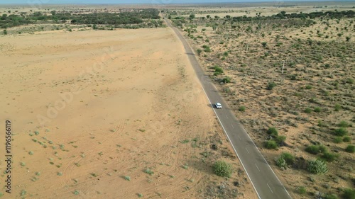A drone shot of a moving car on Longewala Road, Jaisalmer, as it drives through the vast, desert landscape of Rajasthan. The golden sand stretches endlessly beneath a clear blue sky India