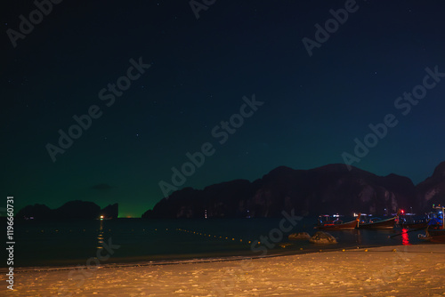 A serene night scene of a sandy beach in Thailand with anchored long tail boats, limestone cliffs silhouetted against a starry sky, and calm waters. photo