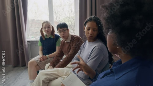 Side view shot of young African American psychologist speaking to diverse zoomers sitting in chairs in circle while leading group therapy session in modern class