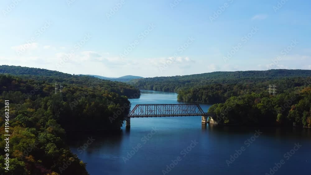 Aerial fly-in towards a steel bridge spanning the New Croton Reservoir, surrounded by lush forested hills in Westchester County, New York, under a clear sky. Aerial fly-in towards a steel bridge spanning the New Croton Reservoir, surrounded by lush forested hills in Westchester County, New York, under a clear sky.