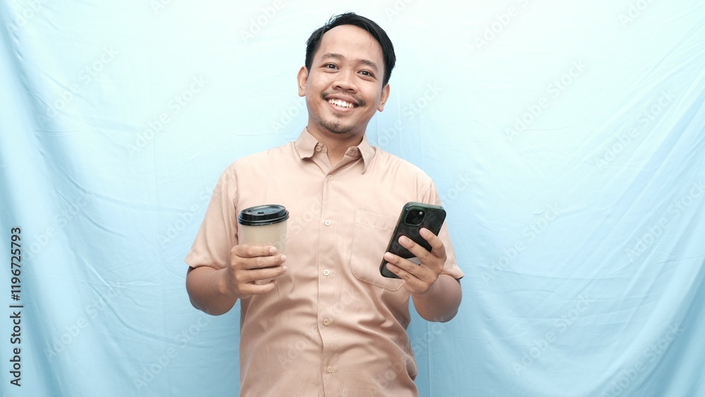 Fototapeta premium Smiling Asian man in a beige shirt holding a coffee cup in one hand and a smartphone in the other, standing against a light blue background.