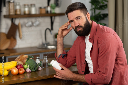 Young man with headache holds a bottle of pills in hands at the kitchen, man with beard taking medicine painkiller medical drug