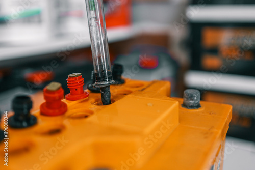 A hydrometer is used to test the fluid density of a yellow car battery. The battery features visible red and black terminals