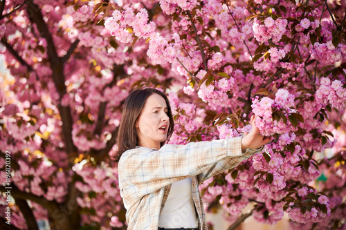Woman allergic suffering from seasonal allergy at spring in blossoming garden at springtime. Young woman having runny nose in front of blooming tree. Spring allergy concept