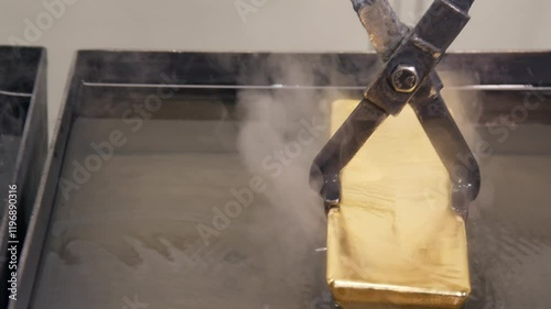 Close-up of a hot gold ingot, moved with tongs from a tank with dark liquid to a tank with a transparent. Process of cooling and hardening of gold after casting is carried out at a metallurgical plant