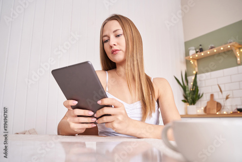 Young caucasian female using tablet in modern kitchen with shelves and plants.