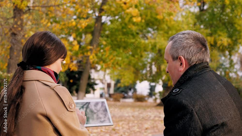 Woman and man have discussion on project looking at tablet. Discussion between man and woman about joint project on tablet with presentation in autumn park. Woman explains project step by step to man