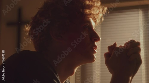 Despaired young man with curly hair praying to God and emotionally crying in warmly-lit room with cross blurred in background