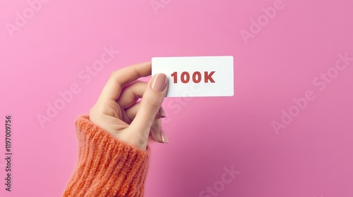 Woman s Hand Holding White Card with 100K Text Against Pink Background Celebrating Online Milestone photo