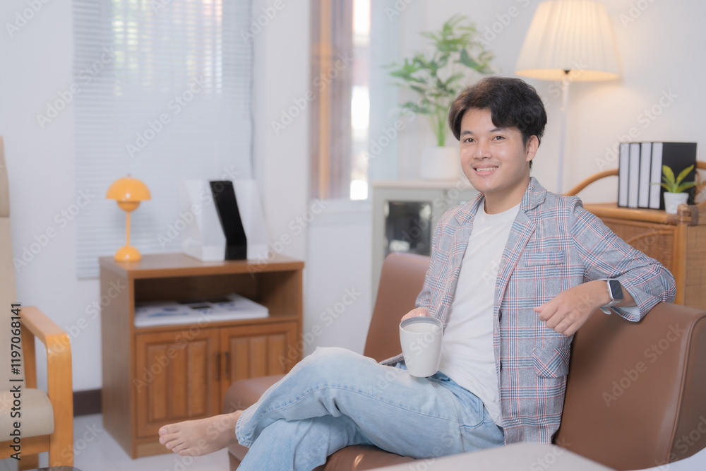 Young Asian businessman wearing casual clothes, sitting on comfortable sofa, drinking coffee and relaxing in living room at home after a long day of work Young Asian businessman wearing casual clothes, sitting on comfortable sofa, drinking coffee and relaxing in living room at home after a long day of work