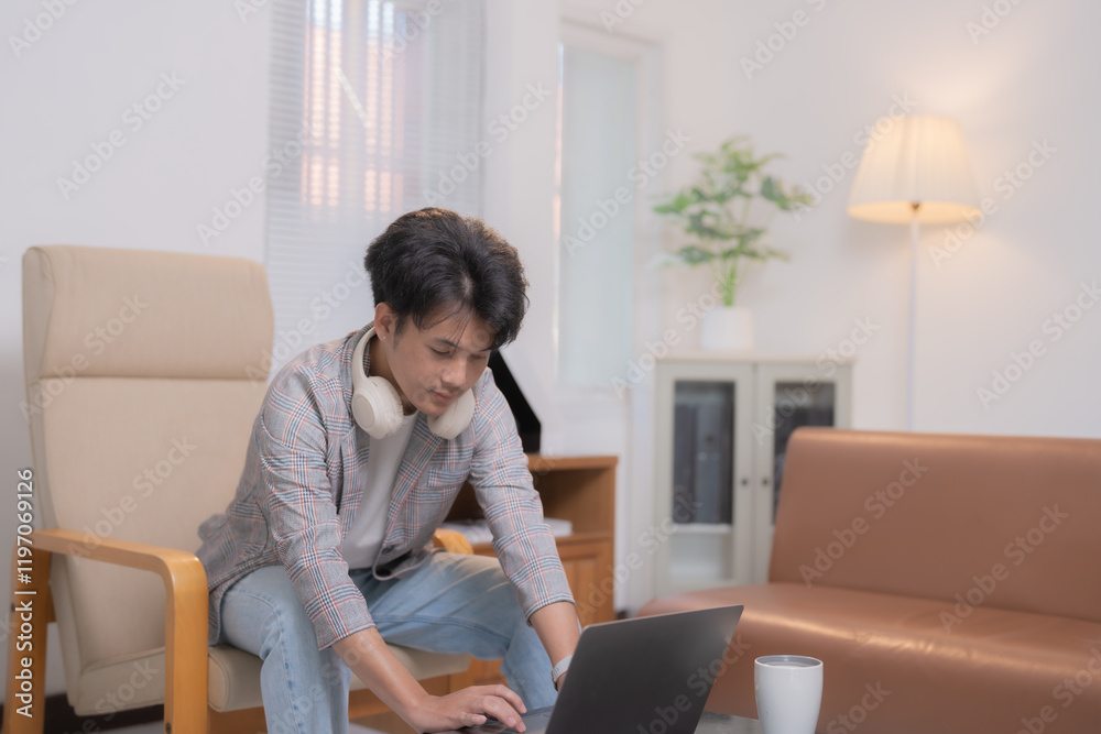 Asian freelancer working from home, using laptop and sitting on armchair, with headphones around his neck, in his living room with sofa, cabinet and lamp Asian freelancer working from home, using laptop and sitting on armchair, with headphones around his neck, in his living room with sofa, cabinet and lamp