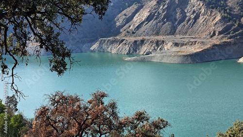 Breathtaking view of a turquoise mountain reservoir nestled between dramatic rocky cliffs. Framed by silhouettes of autumn trees, this stunning landscape captures the perfect harmony of rugged terrain photo