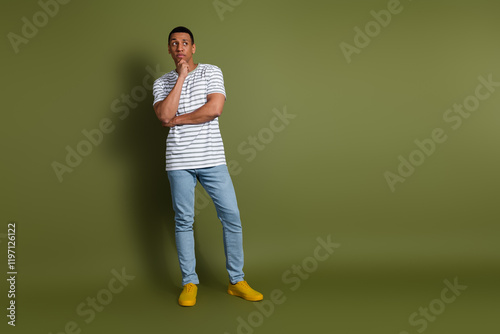 Full length photo of handsome unsure guy wear striped t-shirt arm chin looking emtpy space isolated khaki color background photo