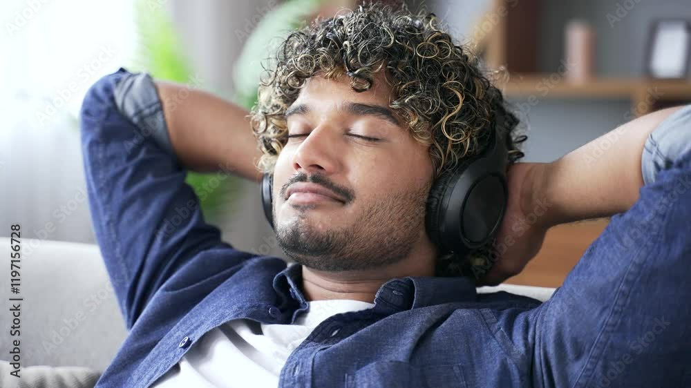 Joyful handsome man wearing wireless headphones enjoys listening music sitting on cozy sofa in living room at home. Happy calm young male relaxes to sounds of relaxation and noise of nature. Close up