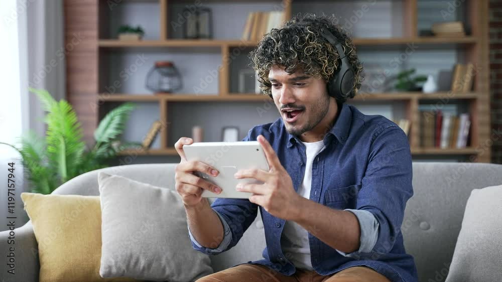 Happy young handsome man in wireless headphones plays a video game on a digital tablet while sitting on sofa in living room at home. Excited concentrated male student engrossed in interesting game Happy young handsome man in wireless headphones plays a video game on a digital tablet while sitting on sofa in living room at home. Excited concentrated male student engrossed in interesting game
