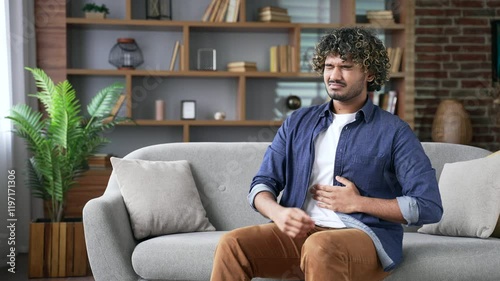 Upset young adult man feels stomach pain while sitting on sofa in living room at home. Sick handsome male has heartburn, gastritis, bloating or poisoning. Male suffers from spasms and constipation