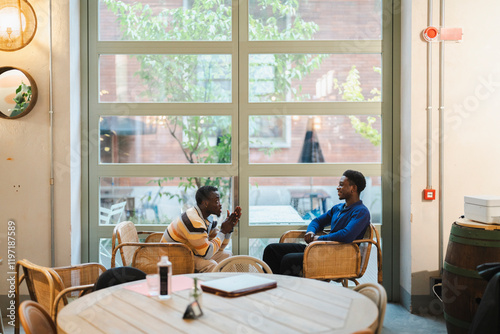 Two black men sitting in a modern cafe environment, deeply engaged in a meaningful conversation while seated in wicker chairs by large windows with natural light. photo