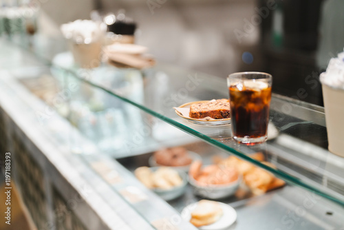 A glass counter in a cafe displays a slice of cake and a glass of iced drink alongside other baked goods, offering a glimpse of the appealing and carefully curated menu items in a modern setting. photo