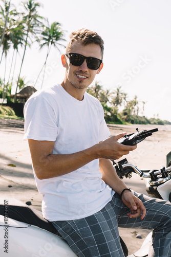 Smiling young trendy biker sitting on motorbike and using uav controller at seaside #1197219107