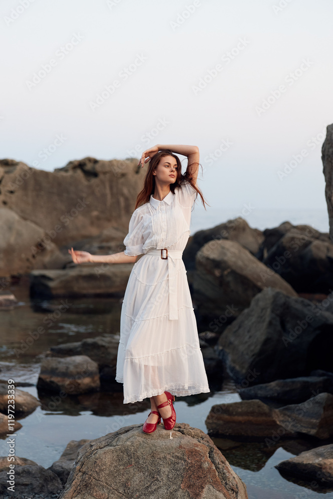 Woman in flowing white dress stands on rocky shore with outstretched arms by the ocean Woman in flowing white dress stands on rocky shore with outstretched arms by the ocean