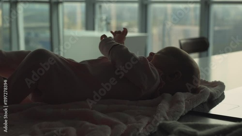 Silhouetted baby rests on blanket while water fills bathtub in background, preparing for swim session with professional instructor. Scene reflects careful preparation for infant swimming photo