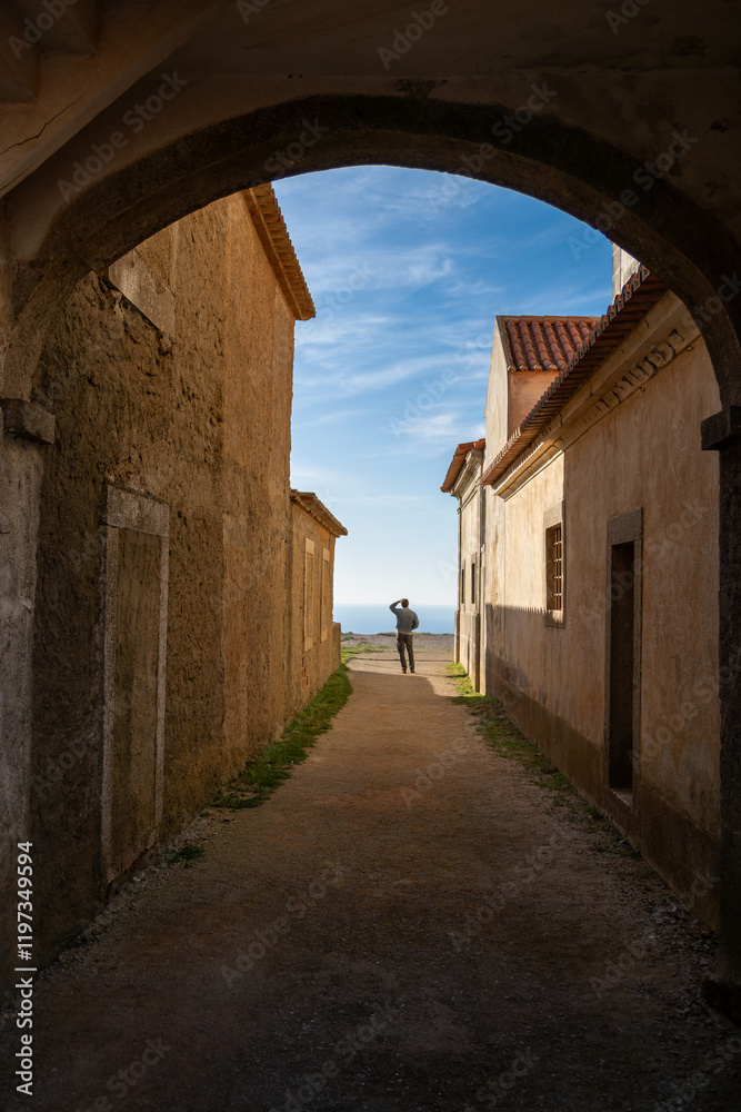 Obraz premium Silhouette of a Man Standing in an Archway Overlooking the Ocean at Santuário de Nossa Senhora do Cabo Espichel, Portugal. December 2024