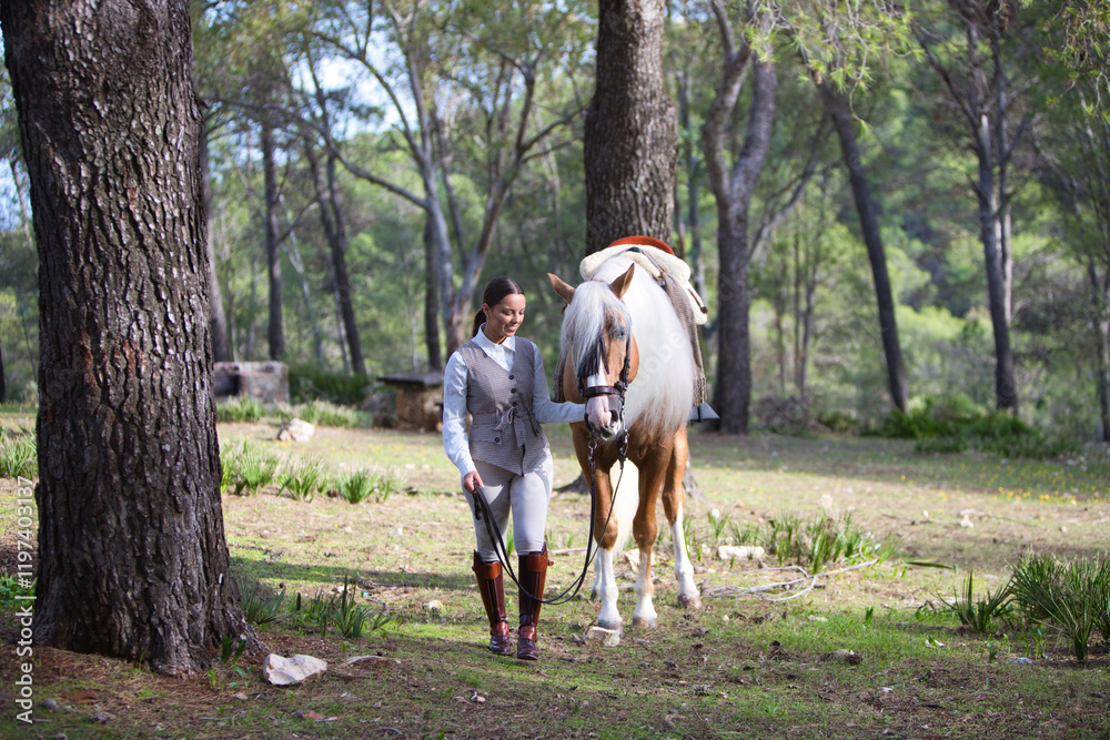 Equestrian woman, young and beautiful, holds the reins and walks through a pine forest with her purebred brown horse with blond mane. The woman is happy. Concept of love for the horse. Equestrian woman, young and beautiful, holds the reins and walks through a pine forest with her purebred brown horse with blond mane. The woman is happy. Concept of love for the horse.