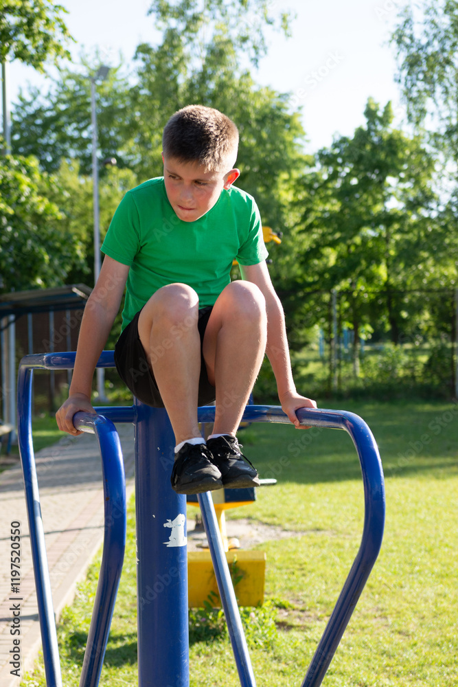 Fototapeta premium A focused young boy performs dips on outdoor fitness equipment. The setting is a sunny park with green grass and trees in the background. He wears a green shirt and dark shorts.
