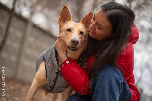 A woman wearing a red jacket and blue jeans is gently holding a dog in a gray vest. They are smiling and interacting affectionately outdoors, showing a loving bond in a natural setting. photo