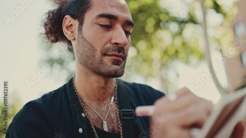 Close-up, young italian guy with ponytail and stubble sits on street bench and makes sketches with pen on sheet of paper on cityscape background