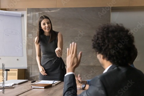 Rear view businessman in suit sits at conference table raising his hand at corporate diverse group meeting, asking question or express opinion at business seminar training led by young female trainer