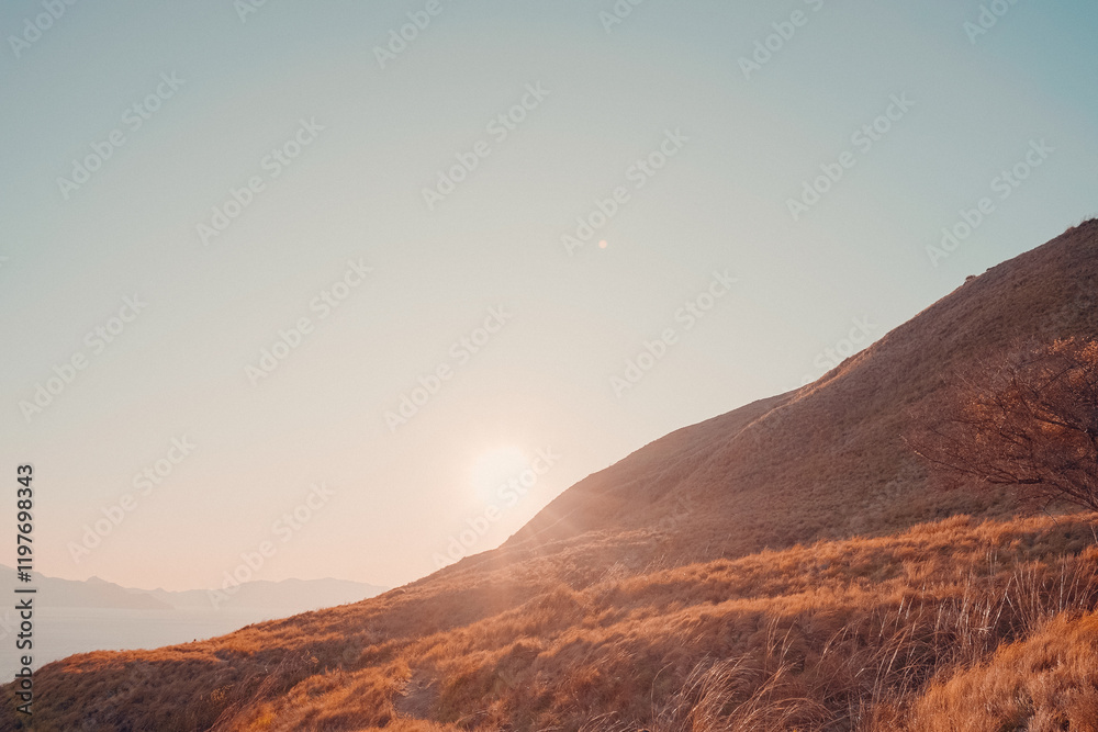 Naklejka premium Stunning panoramic view of Labuan Bajo, Indonesia, featuring golden rolling hills, a tranquil blue ocean with anchored sailboats, and a vibrant sunset glow. 