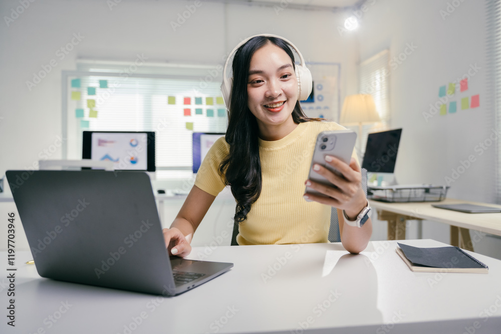 Young asian woman is using laptop and smartphone while working remotely from her home office. She is wearing headphones and smiling Young asian woman is using laptop and smartphone while working remotely from her home office. She is wearing headphones and smiling