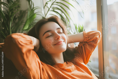 A young woman is resting on the sofa in her living room, smiling and holding her hands behind her head with her eyes closed. photo