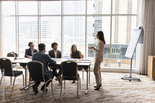 In modern skyscraper office boardroom group of multiethnic businesspeople listening speech of young female Asian corporate coach or trainer, provide information, share strategy. Mentorship, training