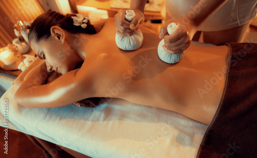 Hot herbal ball spa massage body treatment, masseur gently compresses herb bag on woman body. Tranquil and serenity of aromatherapy recreation in warm lighting of candles at spa salon. Quiescent photo