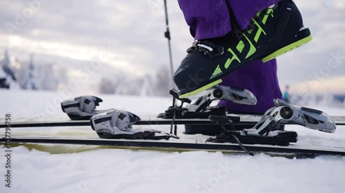 Close-up of skier's legs wearing purple ski pants and black and green ski boots, fastening and unfastening ski bindings on snowy mountain top