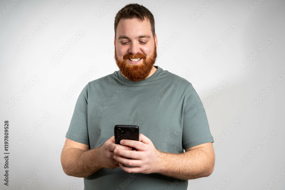 Young happy man looking at his phone, smiling with excitement, standing in a casual outfit against a bright background, enjoying a positive moment while texting or browsing. Young happy man looking at his phone, smiling with excitement, standing in a casual outfit against a bright background, enjoying a positive moment while texting or browsing.