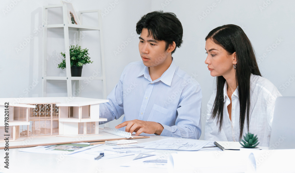 Professional male asian architect using ruler to measure house model length while young beautiful caucasian colleague using laptop to analyzed data on meeting table with house model. Immaculate. Professional male asian architect using ruler to measure house model length while young beautiful caucasian colleague using laptop to analyzed data on meeting table with house model. Immaculate.